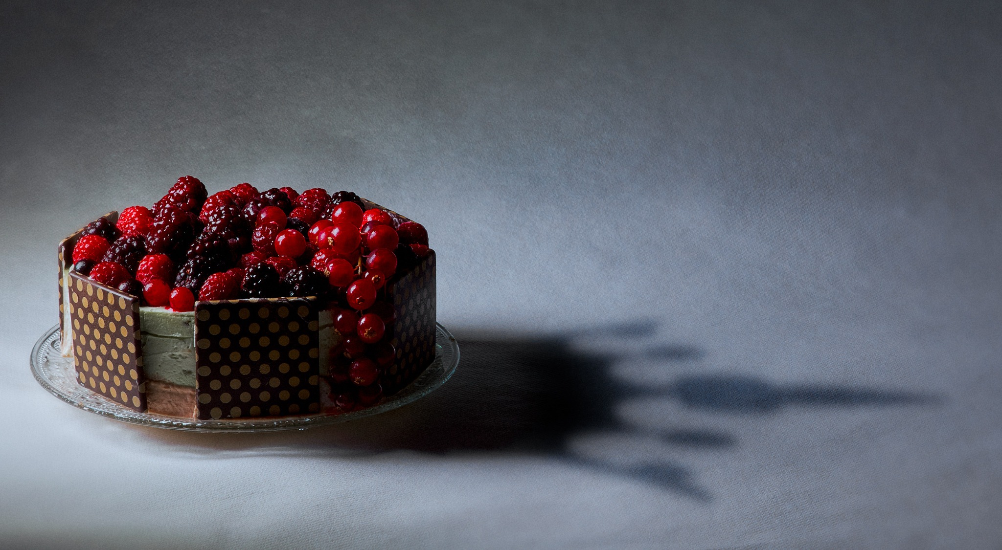 Gâteau au chocolat garni de framboises, mûres et groseilles, photographié sur fond gris.