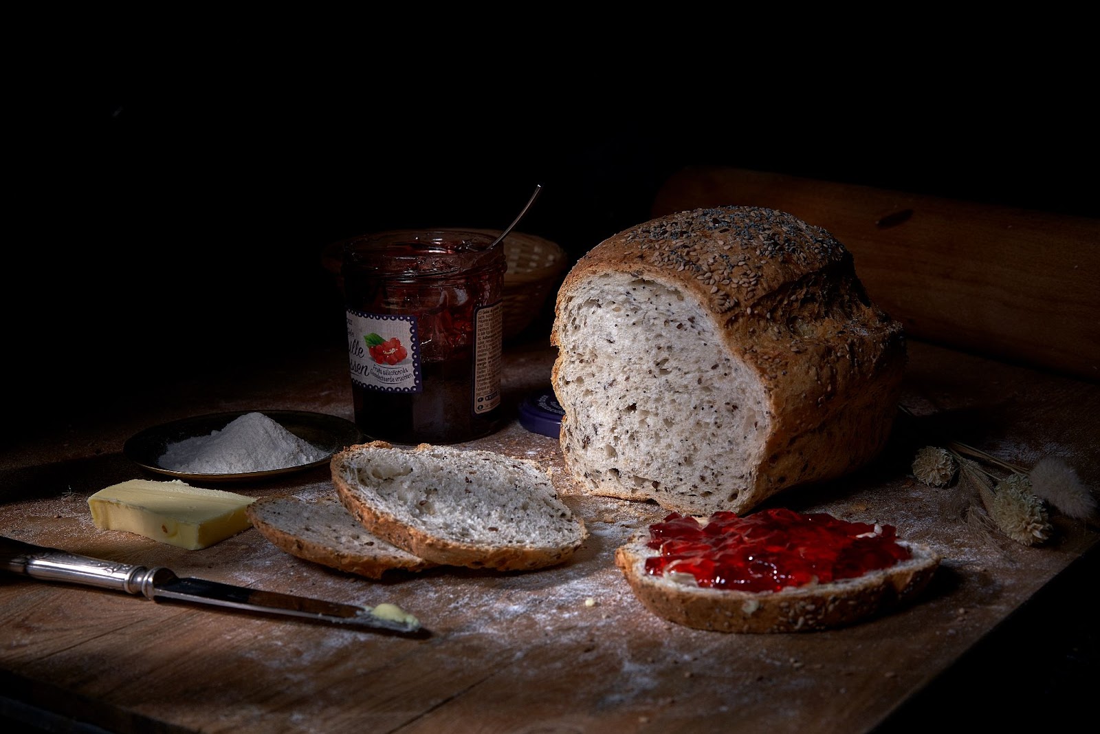 Pain aux graines, tranches beurrées et confiture de fruits rouges sur une table sombre.